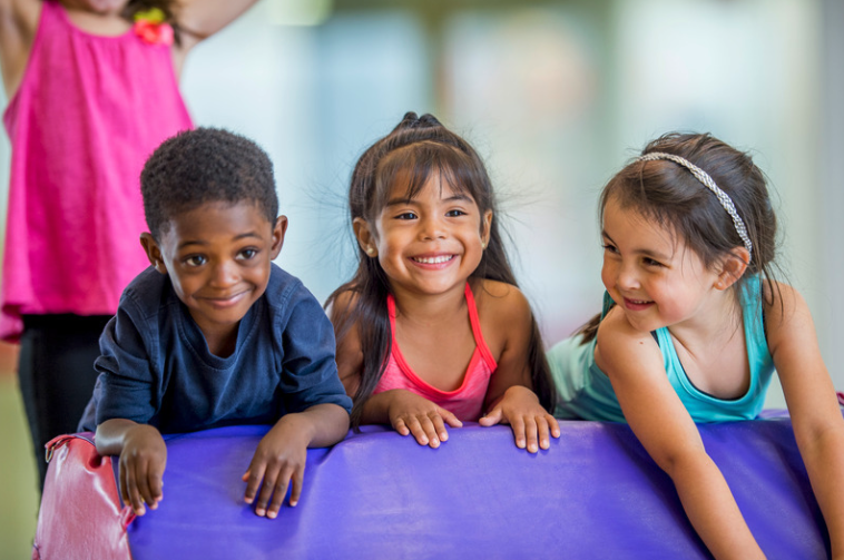Three preschool aged kids on gymnastics equipment smiling. 